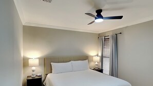 Un dormitorio con una cama, dos mesitas de noche, un ventilador de techo y una ventana con cortinas.