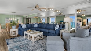 A spacious living room with a sectional sofa, a coffee table, and a patterned rug. There are ceiling fans and a large window providing a view of the outdoors.