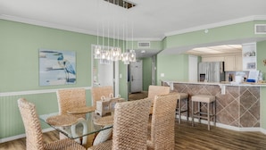 A dining area with wicker chairs and a glass table, a kitchen with a bar, and a painting on the wall.