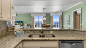 A kitchen with a granite countertop, stainless steel sink, and a dishwasher. There is a view of the ocean through large windows.