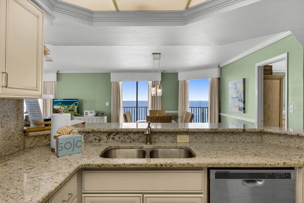 A kitchen with a granite countertop, stainless steel sink, and a dishwasher. There is a view of the ocean through large windows.