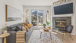 A modern living room with a grey sofa, a fireplace, a flat-screen TV, and a view of mountains through large glass doors.
