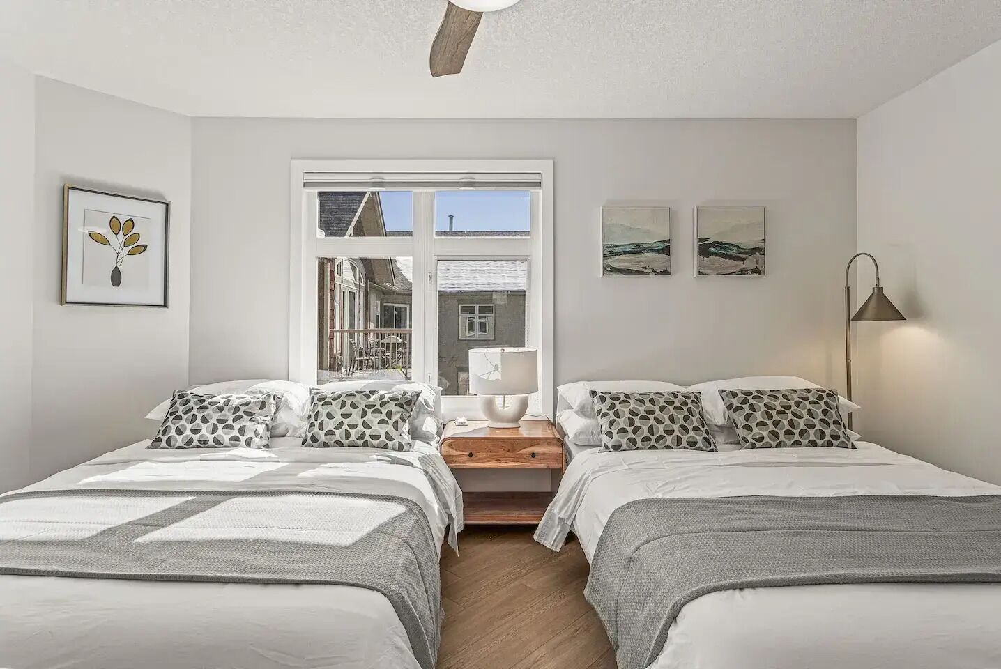 A bedroom with two beds, a window with a view of buildings, a bedside table with a lamp, and wall art.