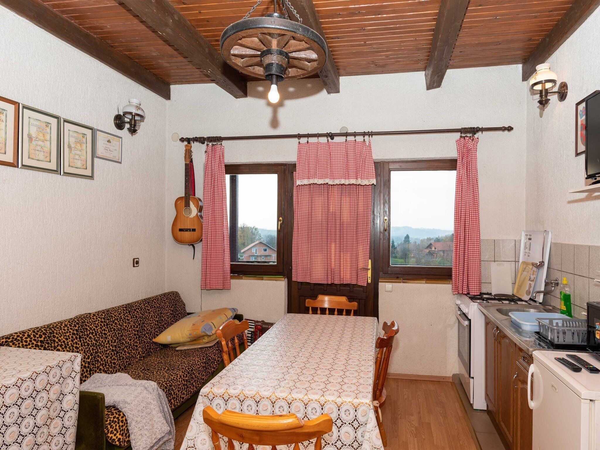 A cozy room with a wooden ceiling, a dining table with chairs, a sofa, a kitchen area with a stove, and a window with red curtains.