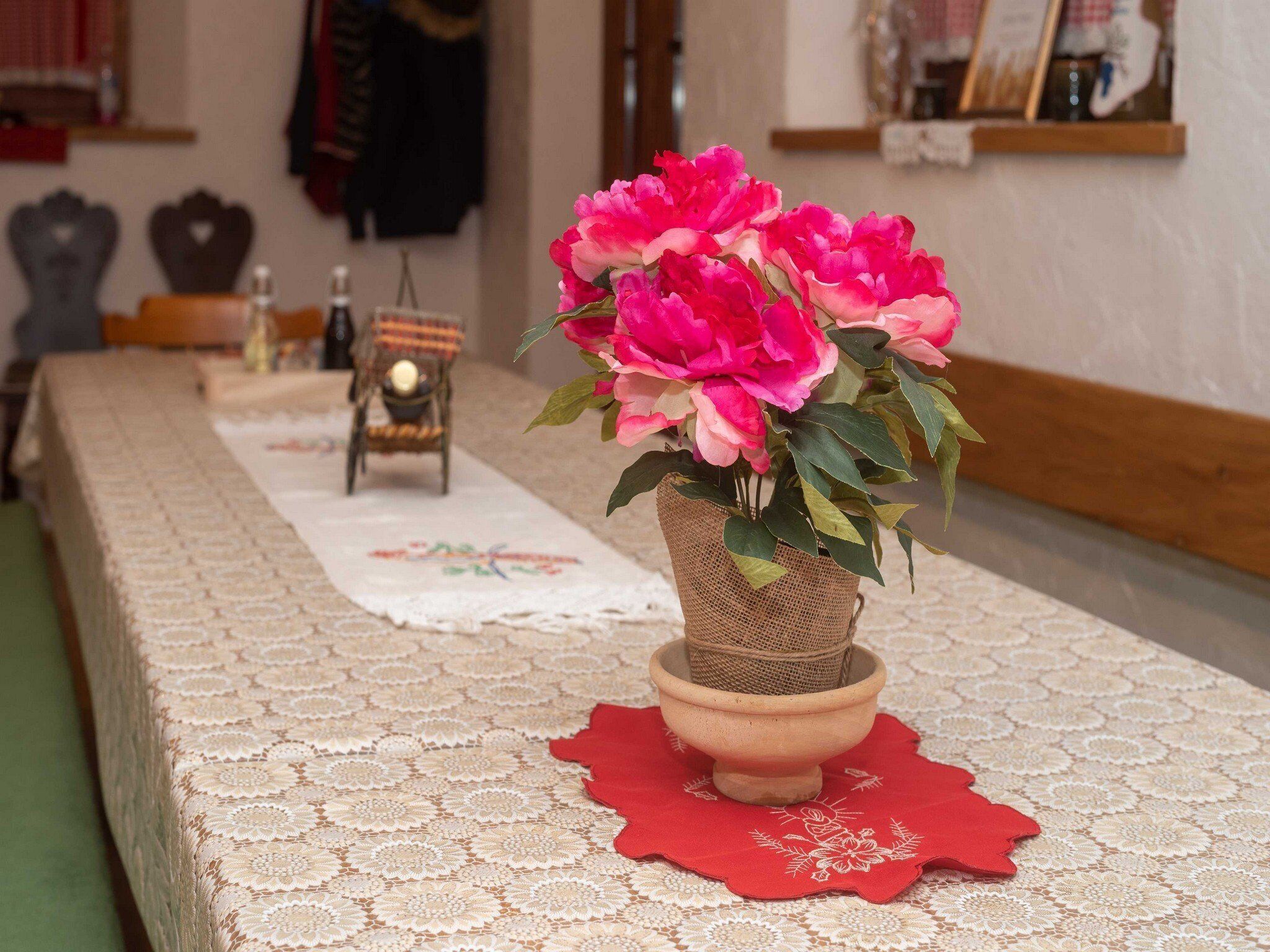 Una mesa con mantel de encaje, camino rojo y una planta en maceta con flores rosas.