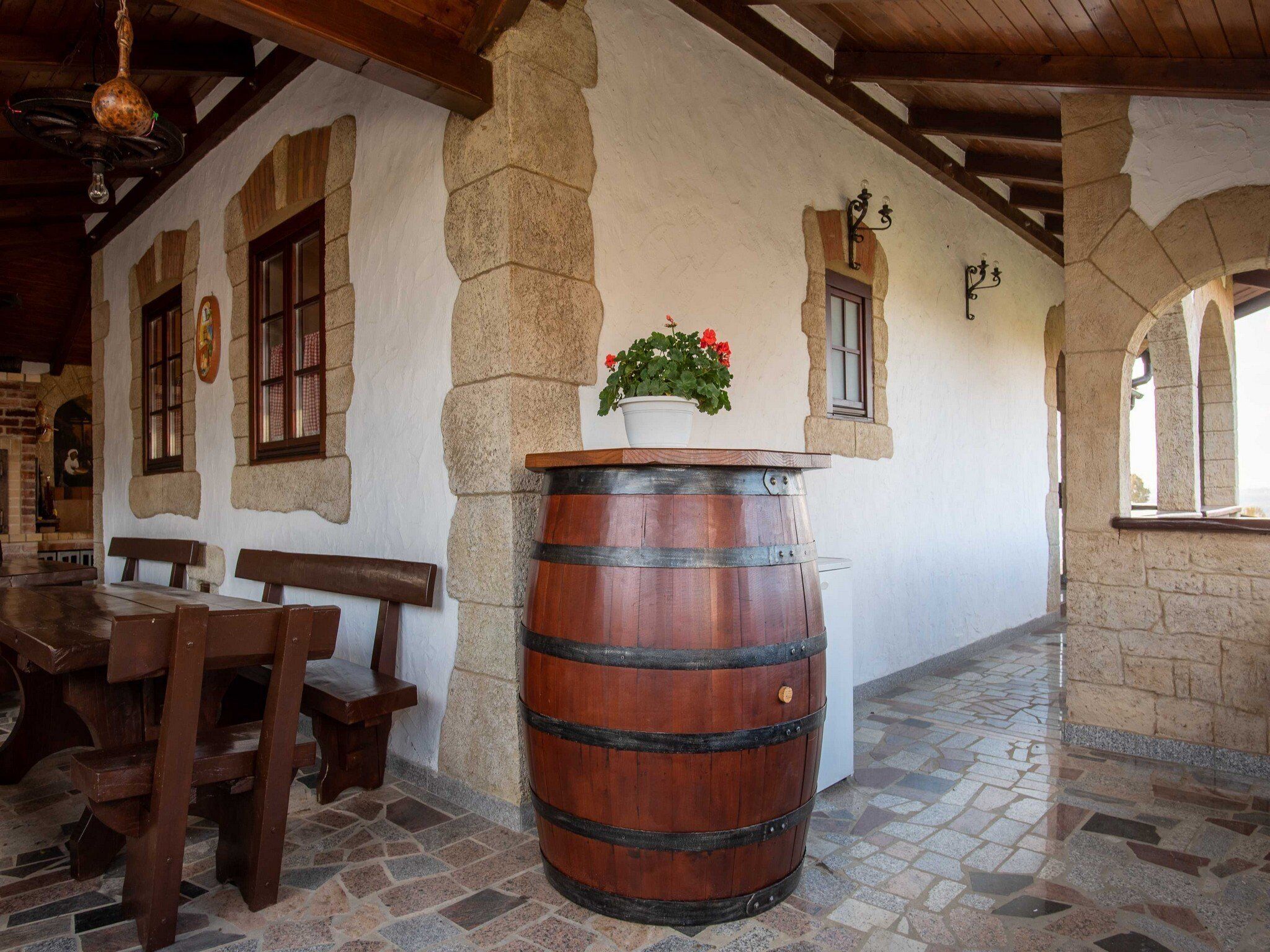Un interior rústico con paredes de piedra, bancos de madera, un barril grande y una planta en maceta.