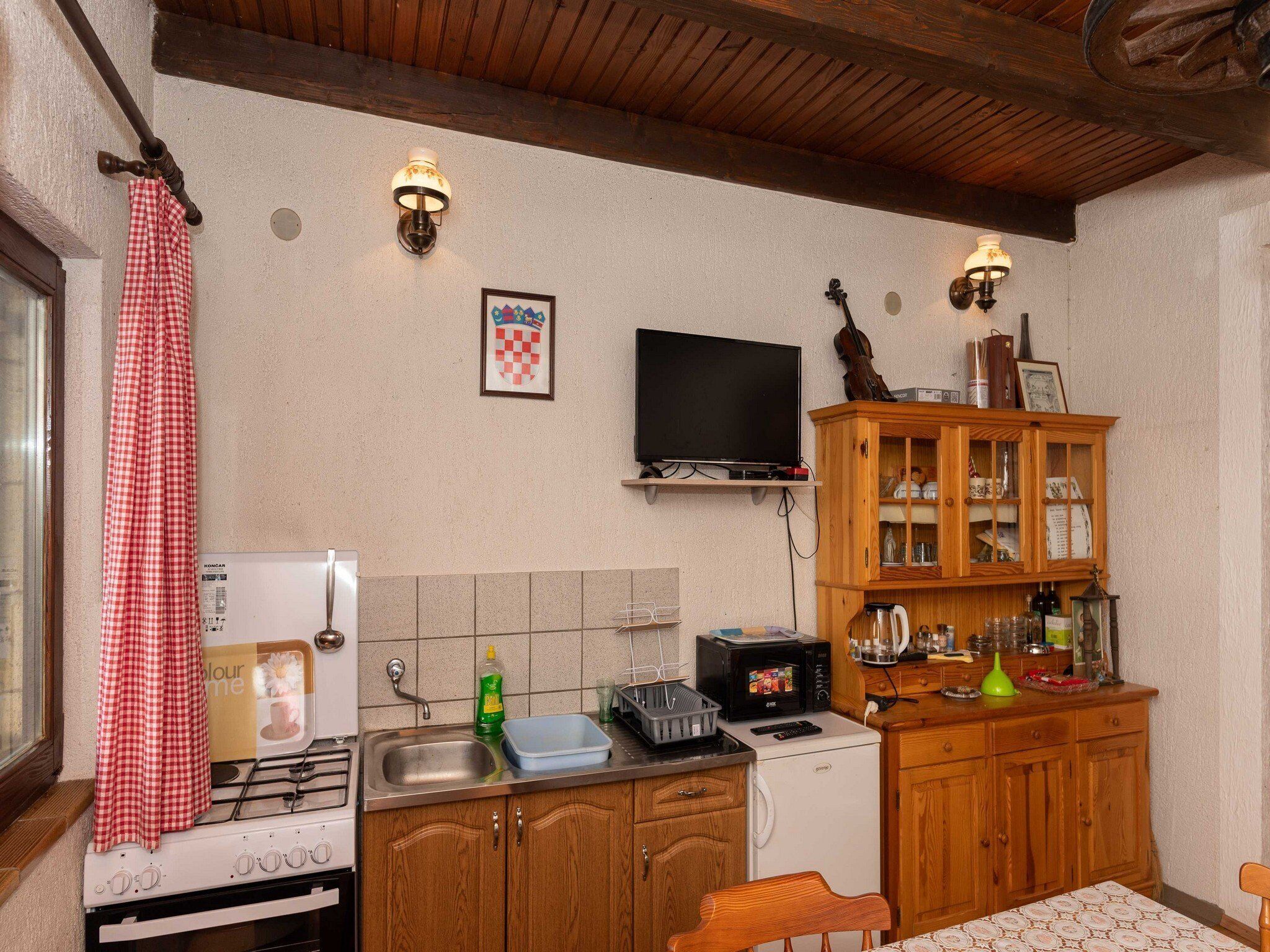 A kitchen with a stove, sink, microwave, and wooden cabinets. There is a television mounted on the wall and a window with a curtain.