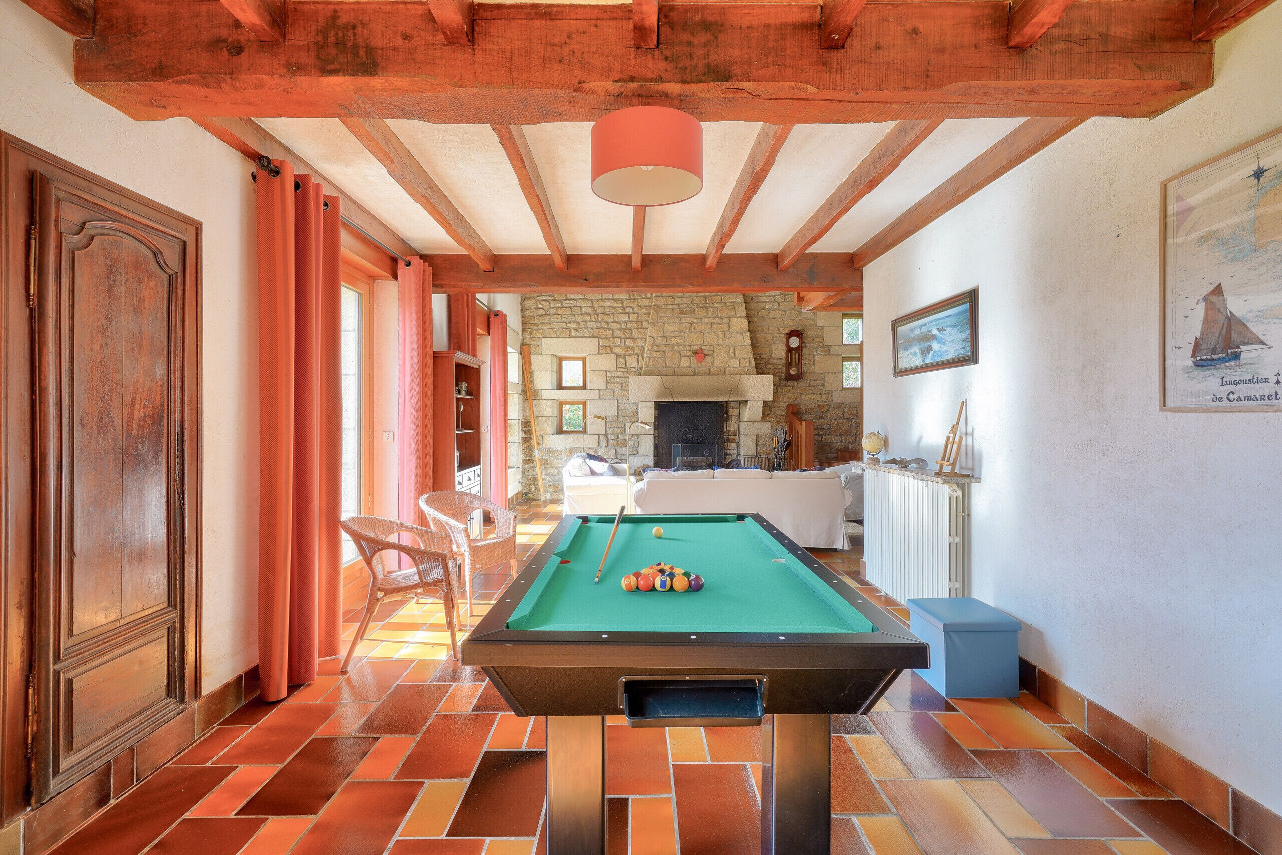 Une table de billard dans une pièce dotée d’une cheminée en pierre, de poutres en bois et d’une grande fenêtre.