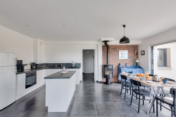 A modern kitchen with a central island, a dining area with a table and chairs, and a brick wall with a wood-burning stove.