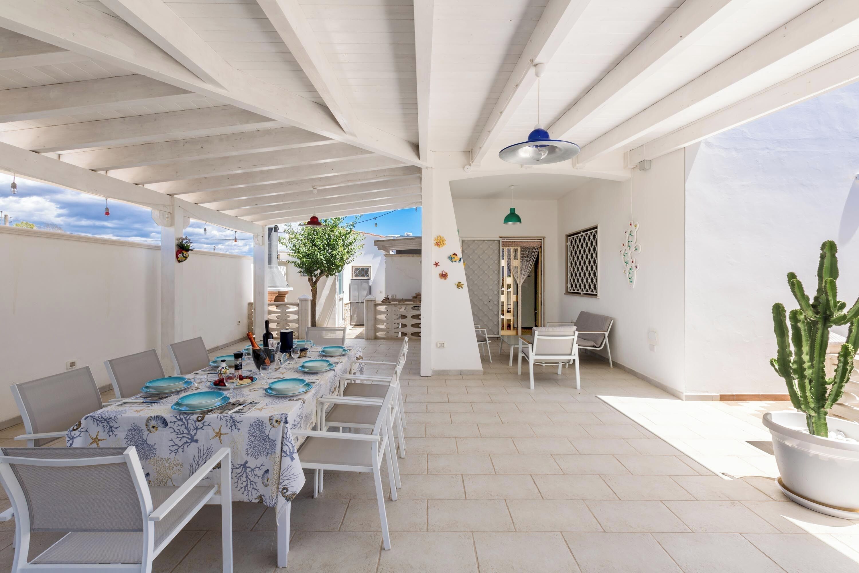 Un espace extérieur abrité, aménagé pour les repas, avec une table pour quatre personnes, des chaises blanches, un cactus en pot et une vue sur le ciel.