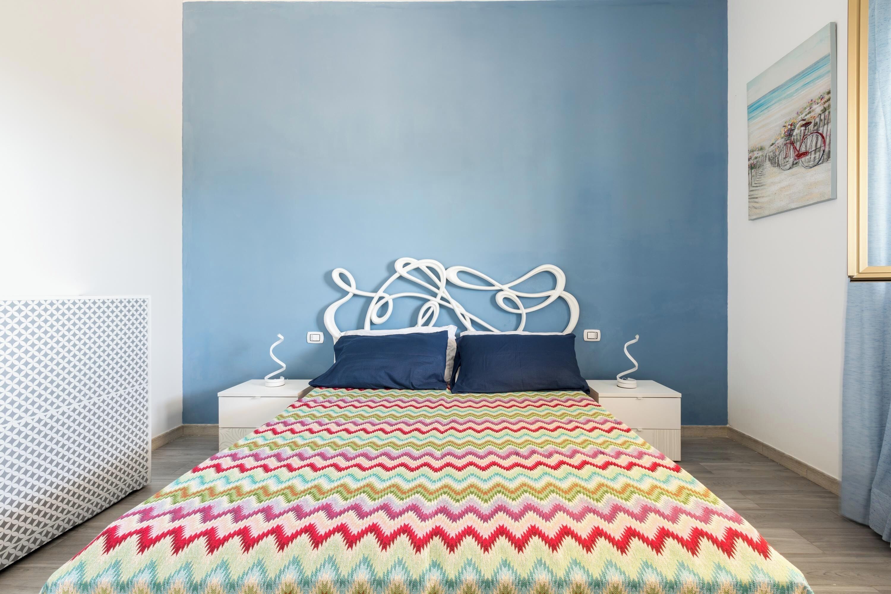 Une chambre à coucher avec un lit, deux tables de chevet, une couvre-lit à motifs et un mur orné d’un tableau.