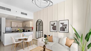 A modern living room with a white sofa, a coffee table, and a dining area with chairs. There is a kitchen in the background. - Silkhaus Uniestate Sports Tower - DSC (Dubai)