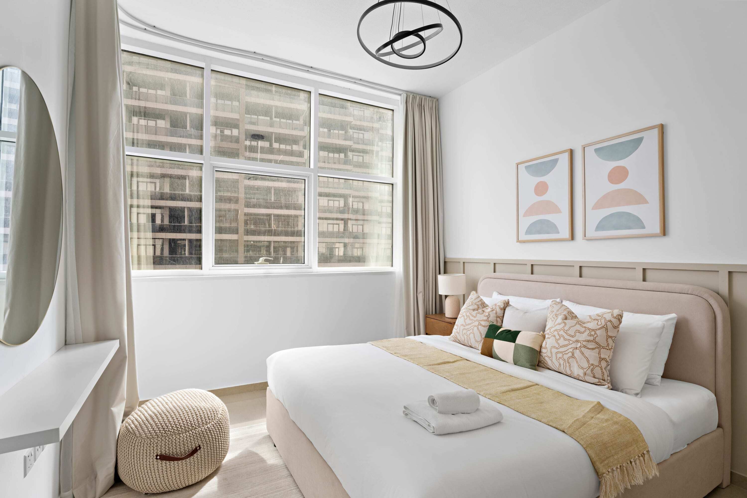 A modern hotel room with a large window, a bed with multiple pillows, a bedside table with a lamp, and a wall-mounted artwork.