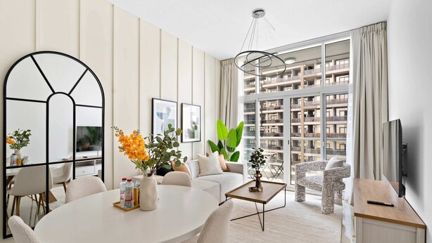 A modern living room with a round dining table, sofas, a coffee table, and a TV. There are large windows with a view of a high-rise building. - Silkhaus Uniestate Sports Tower - DSC (Dubai)