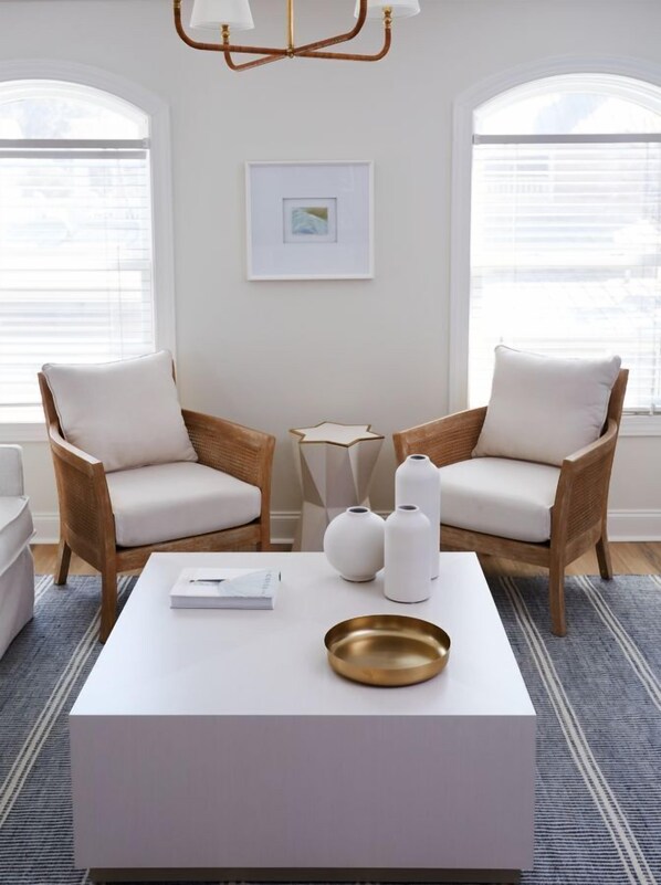 A modern living room with two wicker chairs, a white coffee table, and a gold bowl. There is a framed picture on the wall and a window with blinds.