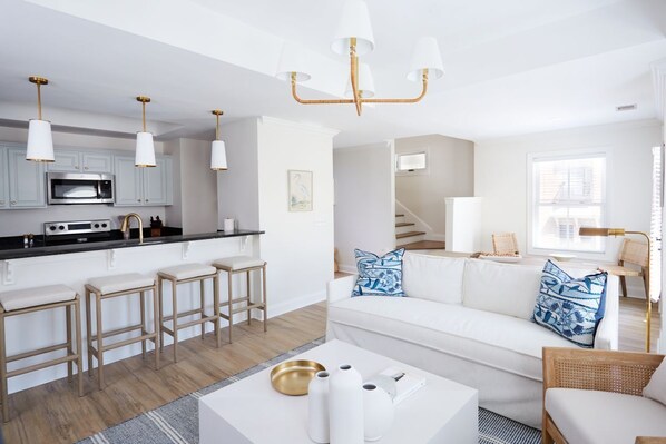 A modern kitchen with white cabinets, a microwave, and bar stools. There is a white sofa with patterned cushions in the living area, and a staircase leading to another level.