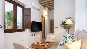 Un salon avec une table basse ronde en bois, des fauteuils beiges, un canapé agrémenté de coussins à motifs floraux, un téléviseur fixé au mur et une lampe.