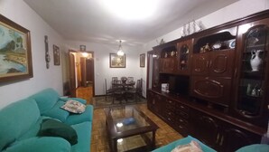 A living room with a teal sofa, a glass coffee table, a wooden cabinet with glass doors, and a dining area with chairs and a table.