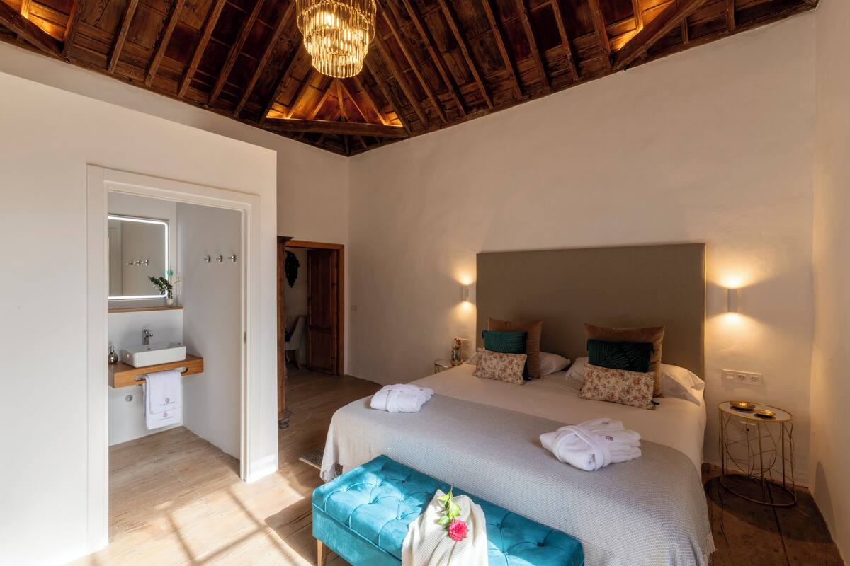 Quarto com uma cama grande, um banco, uma mesa de cabeceira, um espelho e teto de madeira.