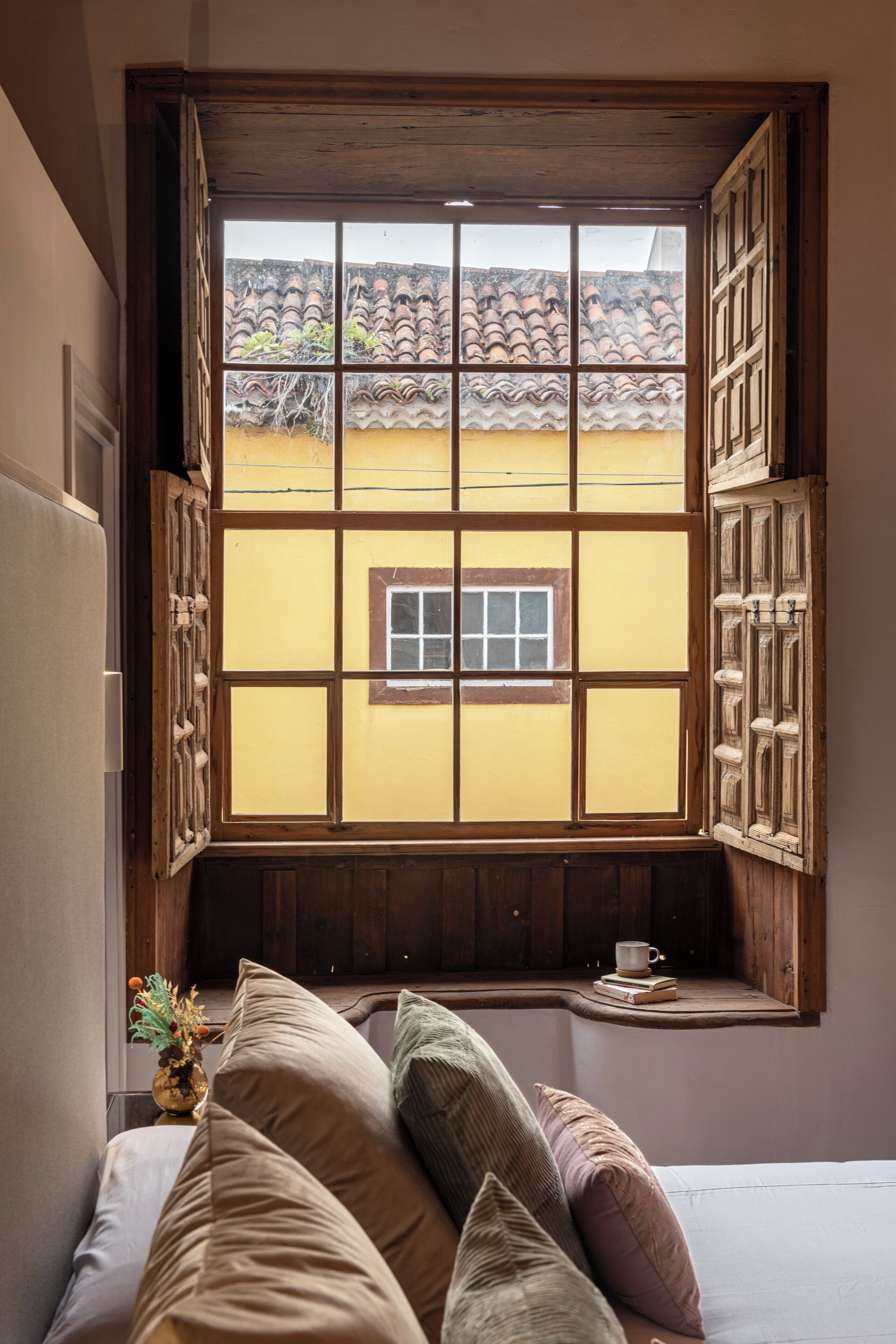 Une pièce dotée d’une grande fenêtre donnant sur un immeuble jaune avec un toit en tuiles. On y trouve un lit avec plusieurs oreillers et une petite table sur laquelle sont posés une tasse et des livres.