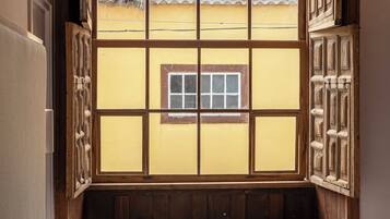 Une pièce dotée d’une grande fenêtre donnant sur un immeuble jaune avec un toit en tuiles. On y trouve un lit avec plusieurs oreillers et une petite table sur laquelle sont posés une tasse et des livres.