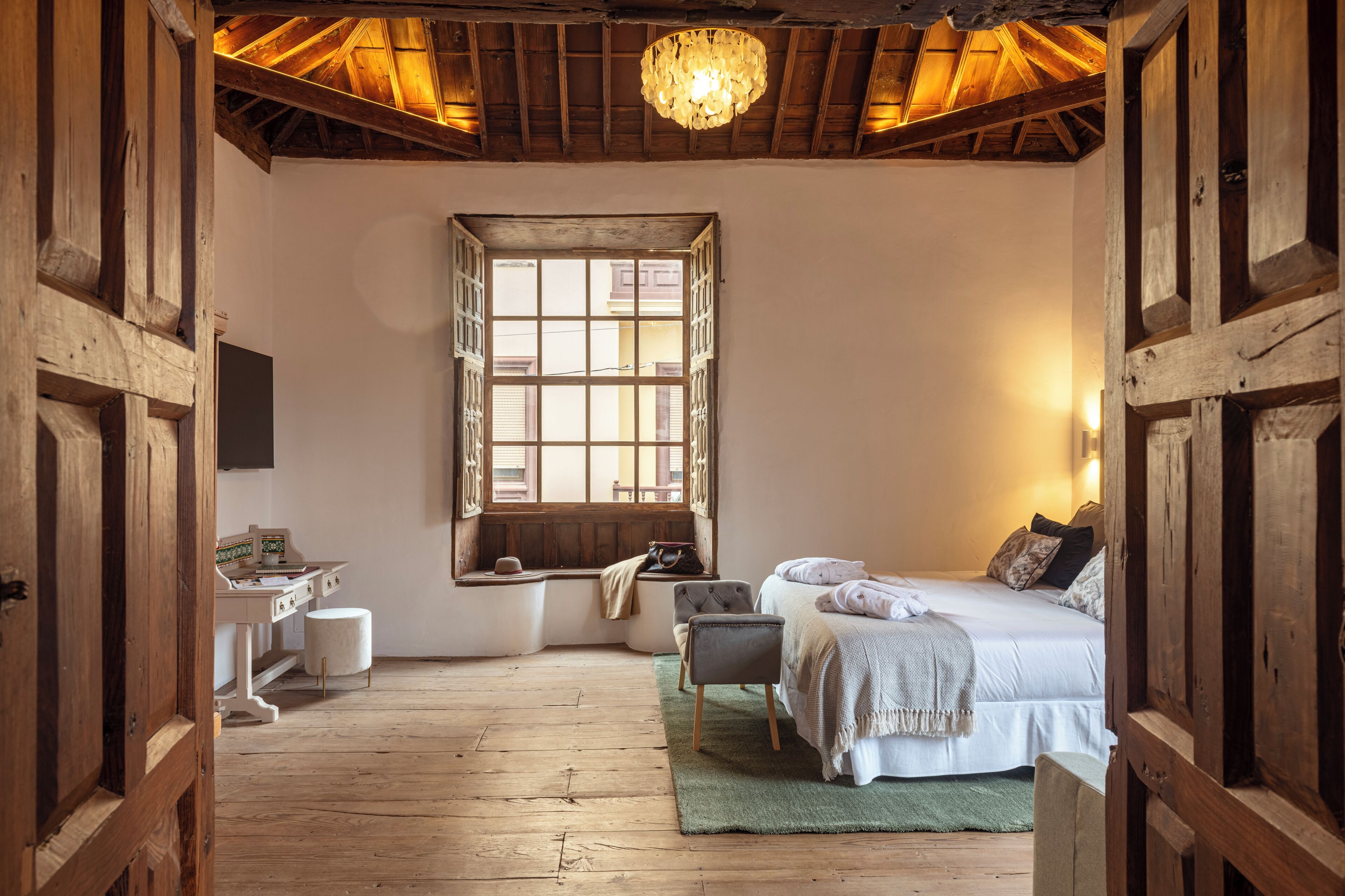 A rustic bedroom with a wooden ceiling, a large window, a bed with pillows, a chair, and a small desk.