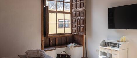 A bedroom with a large window, a bed, a desk with drawers, a chair, and a small trash bin.