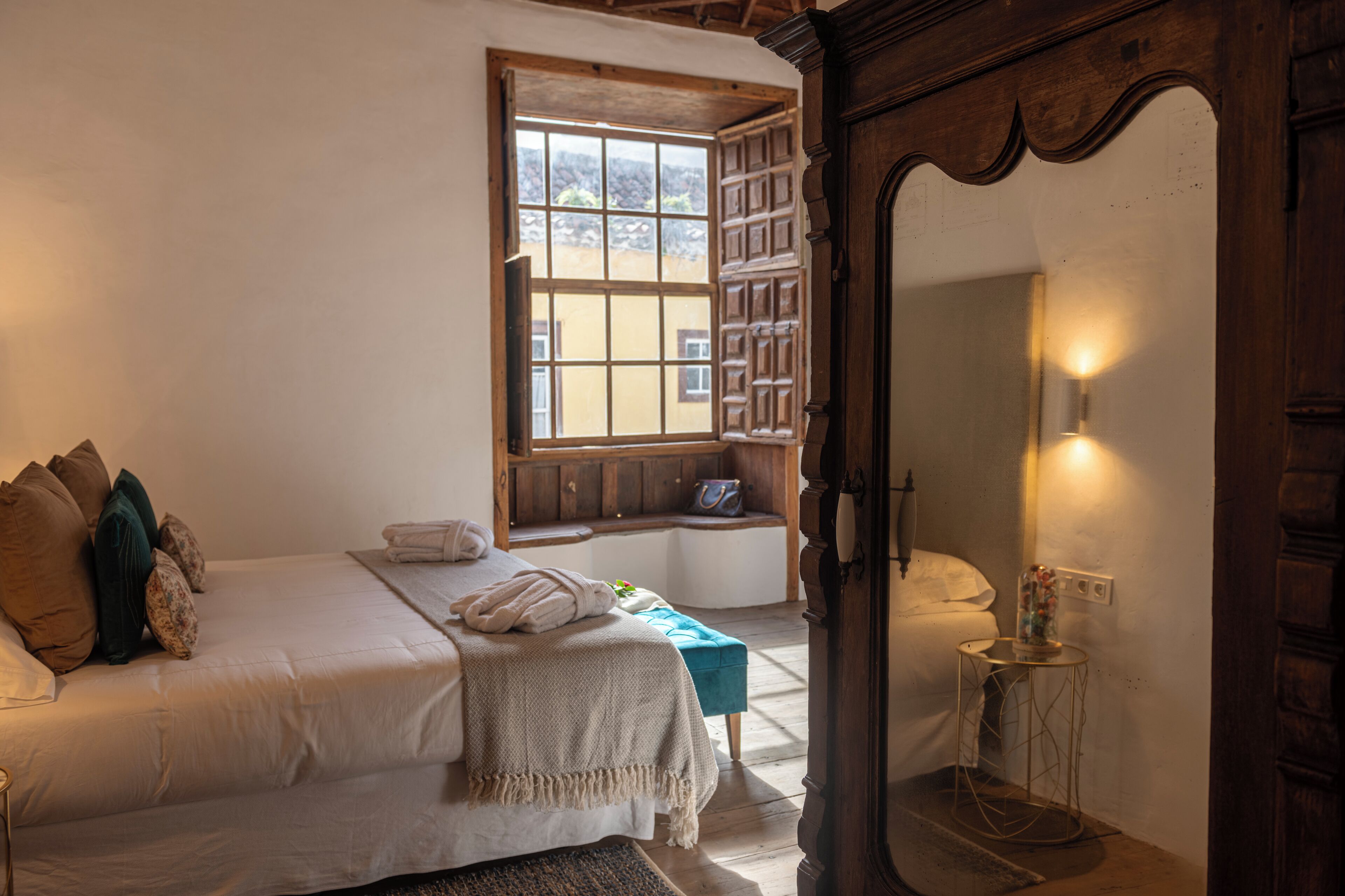 A bedroom with a large window, a bed with pillows, a bench, a small table, and a wardrobe.
