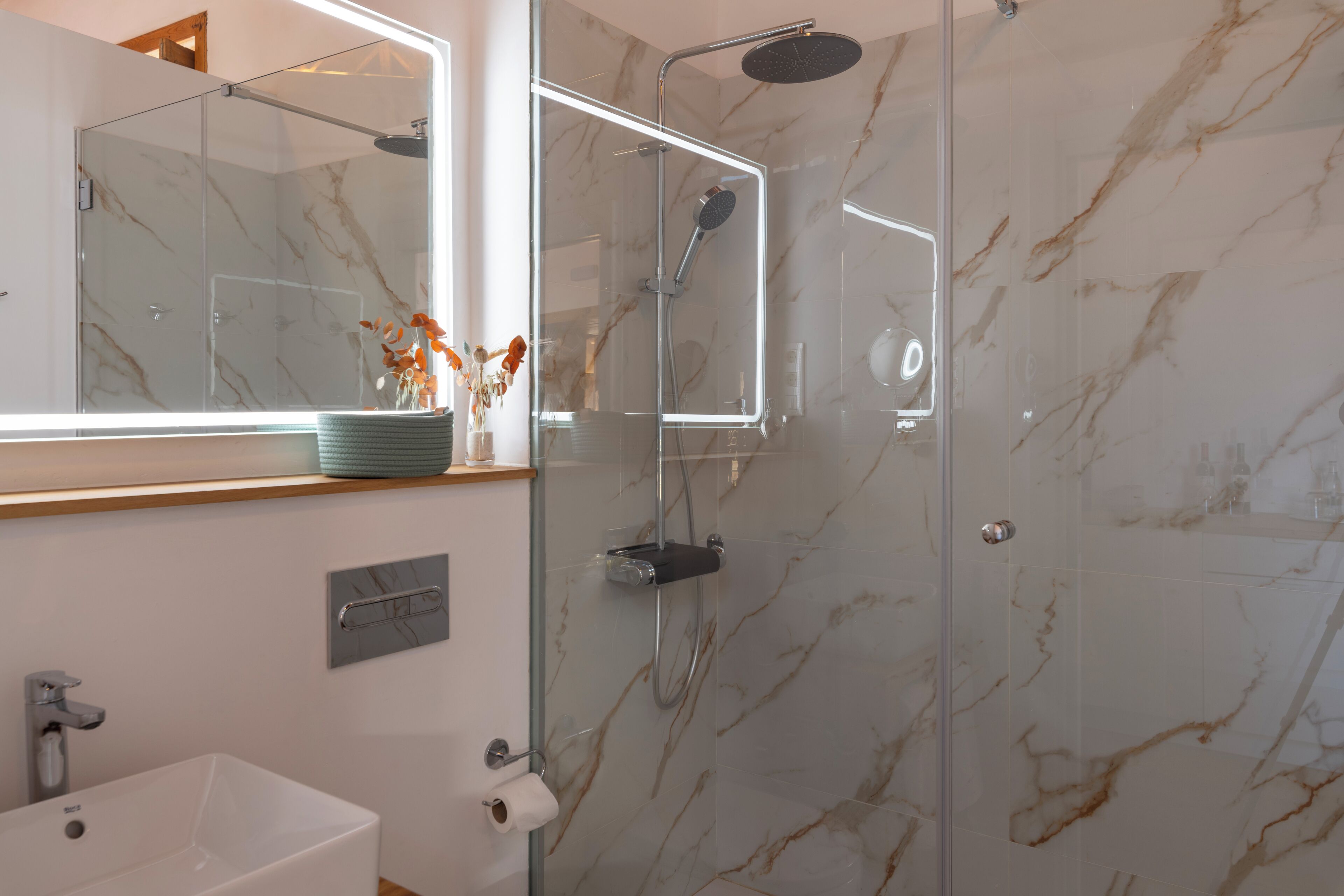 A modern bathroom with a glass-enclosed shower, a marble-tiled wall, a white sink, and a mirror with a light strip.