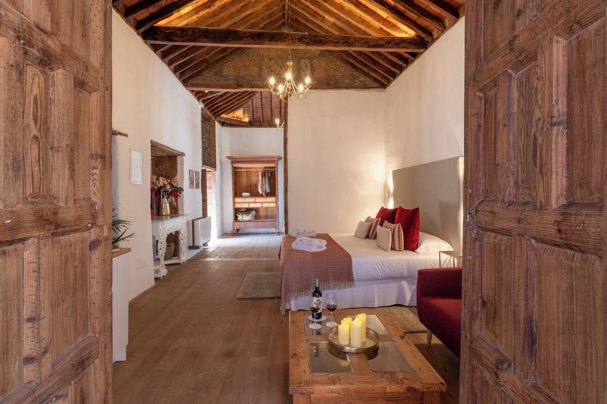 Quarto acolhedor com teto de madeira, uma cama e uma cadeira vermelha, mesa de centro de madeira com velas, e um lustre.