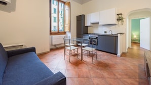 Un espacio habitable compacto con kitchenette, zona de comedor con sillas, sofá y ventana con vista a edificios.