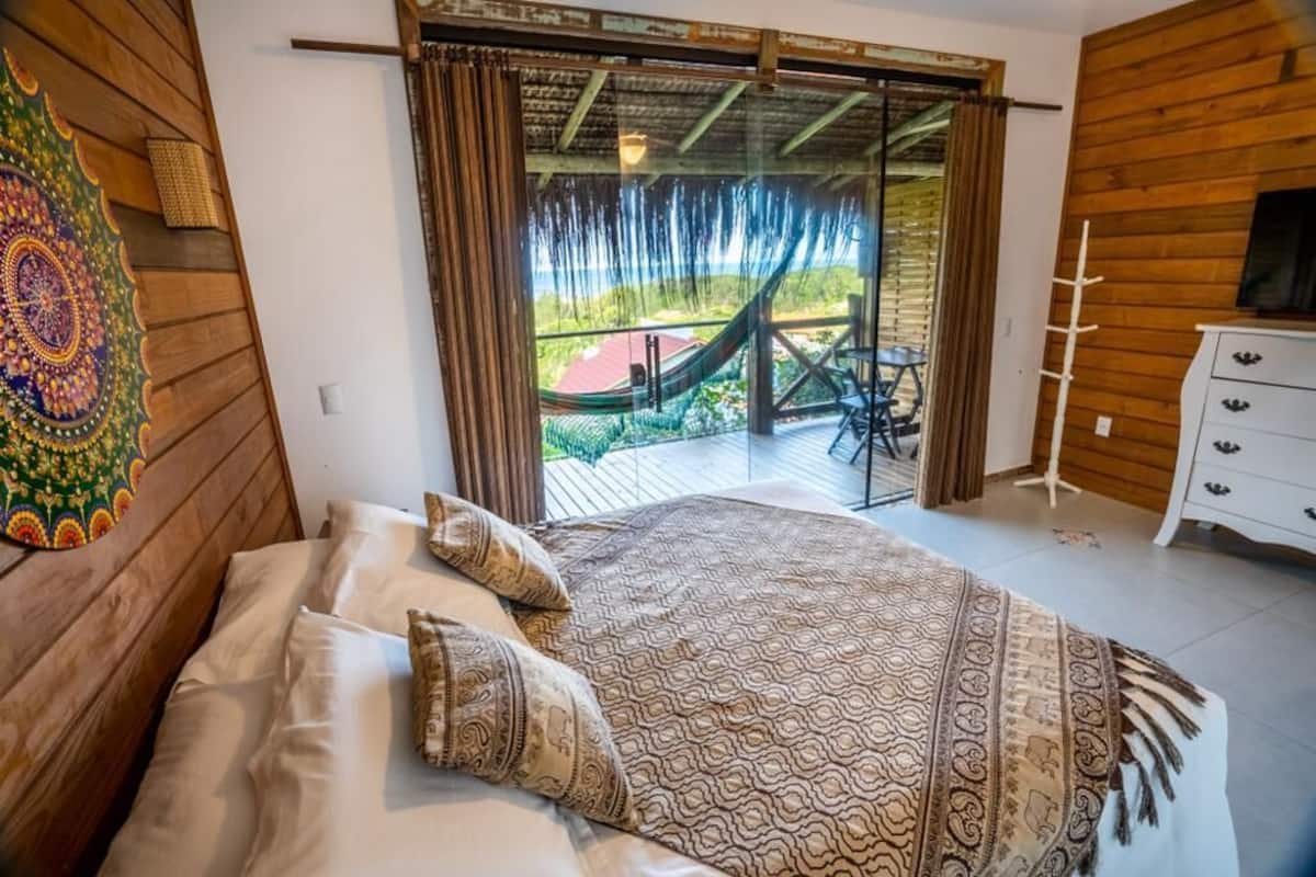 A bedroom with a bed, a wooden headboard, a large window with curtains, a view of a garden, and a small table with chairs outside.