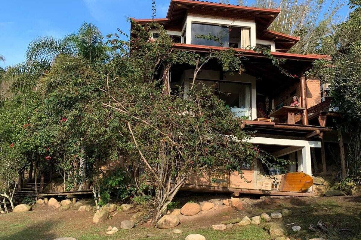 A two-story house with a red roof, surrounded by lush greenery and large rocks.