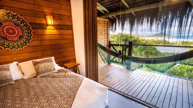 A bedroom with a large bed, a wooden headboard, a wall tapestry, and a view of a hammock and greenery through a glass door. - Cabanas Rosa Mistica (Imbituba)