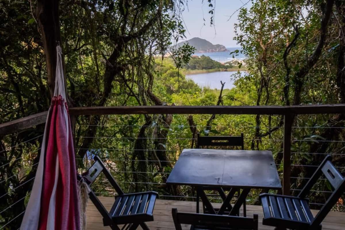 A wooden deck with a table and chairs, overlooking a lake and a mountainous landscape.