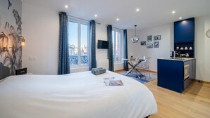 A modern hotel room with a large bed, a small kitchenette, a dining area, and a balcony with curtains.