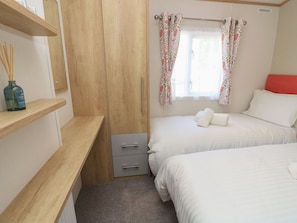 A bedroom with two beds, a wardrobe, a window with floral curtains, and a bedside table with a bottle.
