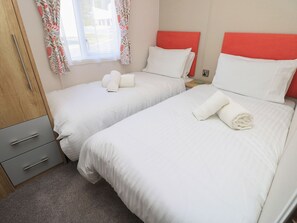 A hotel room with two beds, each with white bedding and red headboards. There are two bedside tables with lamps, a wooden wardrobe, and a window with floral curtains. - 39 Crosswinds (Bembridge)