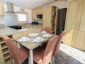 A compact kitchen with a central island, wooden cabinets, and a dining area with pink chairs. - 39 Crosswinds (Bembridge)