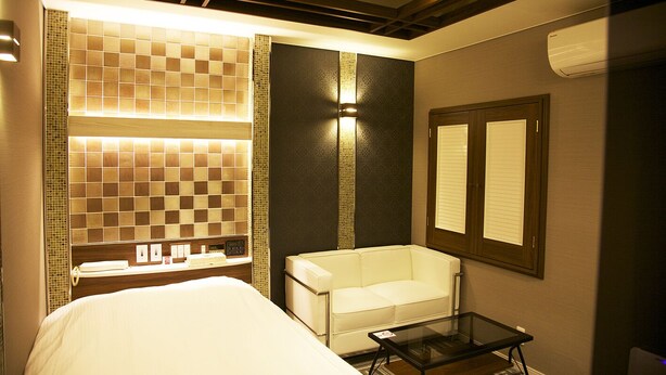 A hotel room with a bed, a small table, a chair, a mirror, and a wall with a patterned tile design. - HOTEL Let's (Saitama)