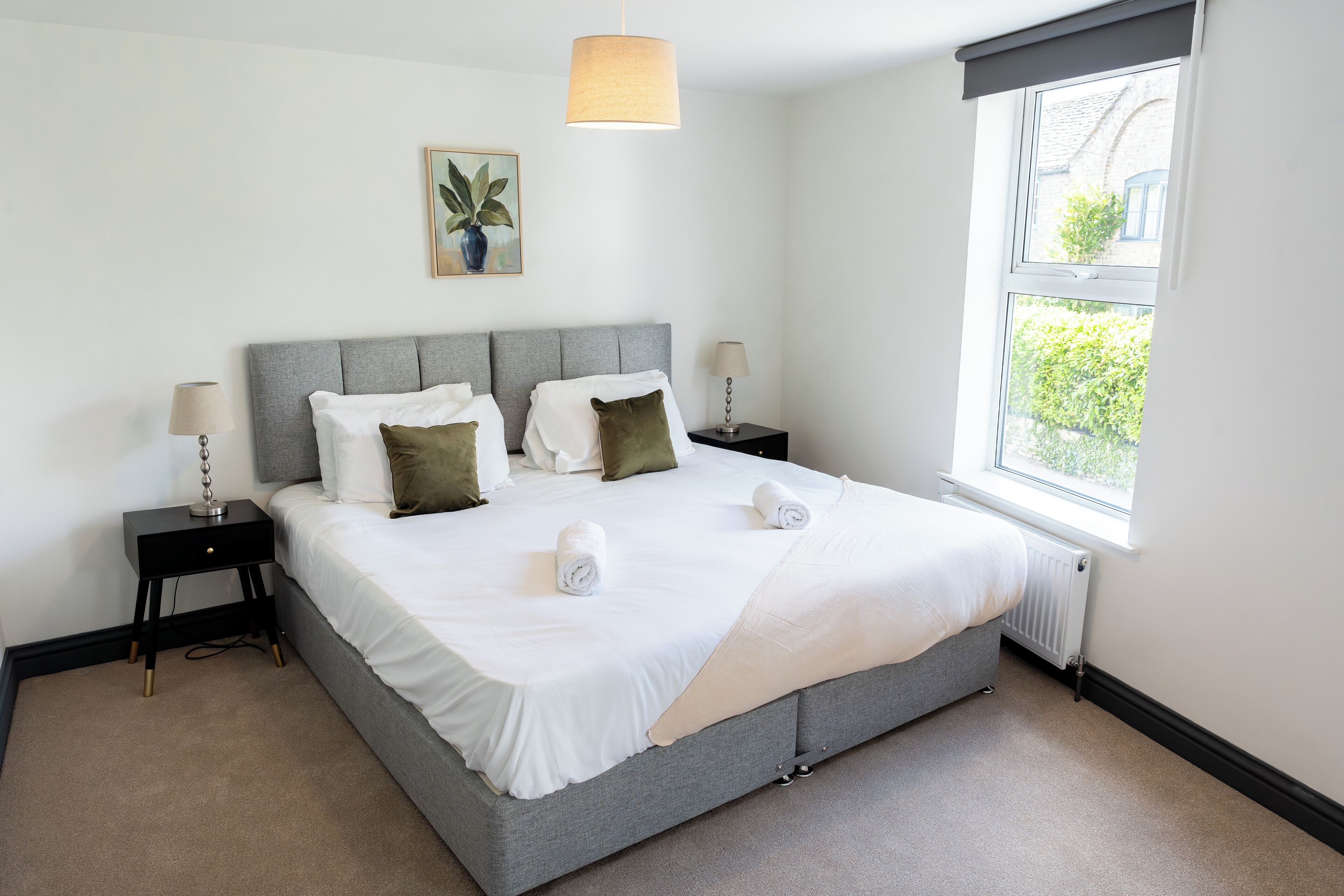A neatly made bed with white linens, two bedside tables with lamps, a grey headboard, and a window with a view of greenery.