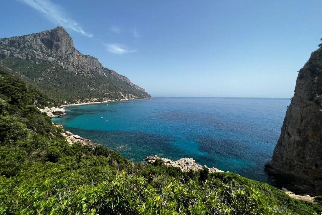 A coastal landscape with a prominent mountain, clear blue sea, and lush greenery.