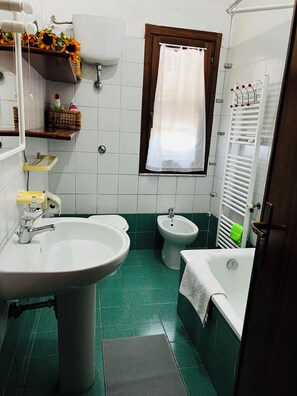 Een badkamer met een wastafel, bidet en badkuip. Er is een raam met gordijn, een handdoekrek en een plank met toiletartikelen.