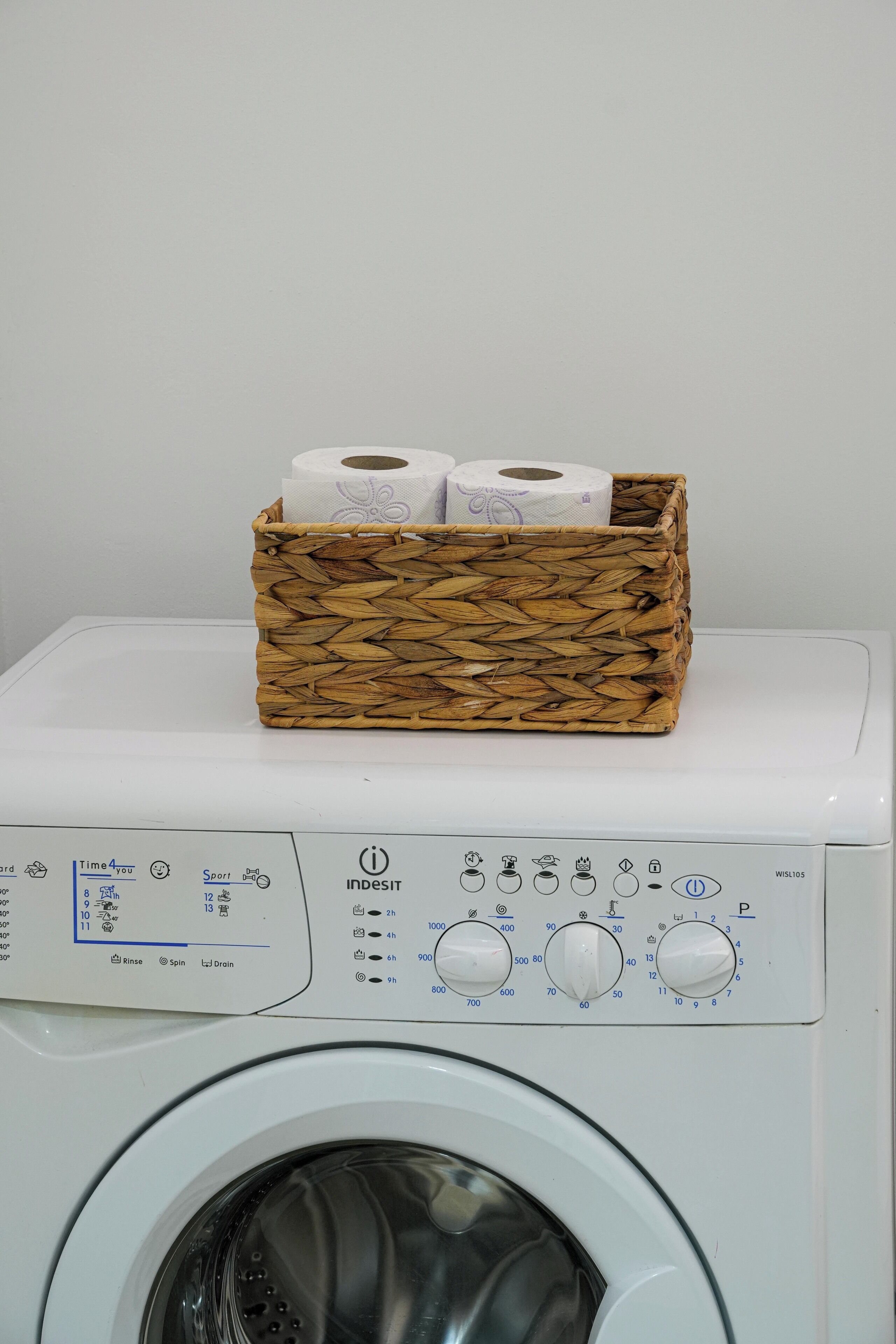 Una lavadora con una cesta de mimbre que contiene dos rollos de papel higiénico encima.