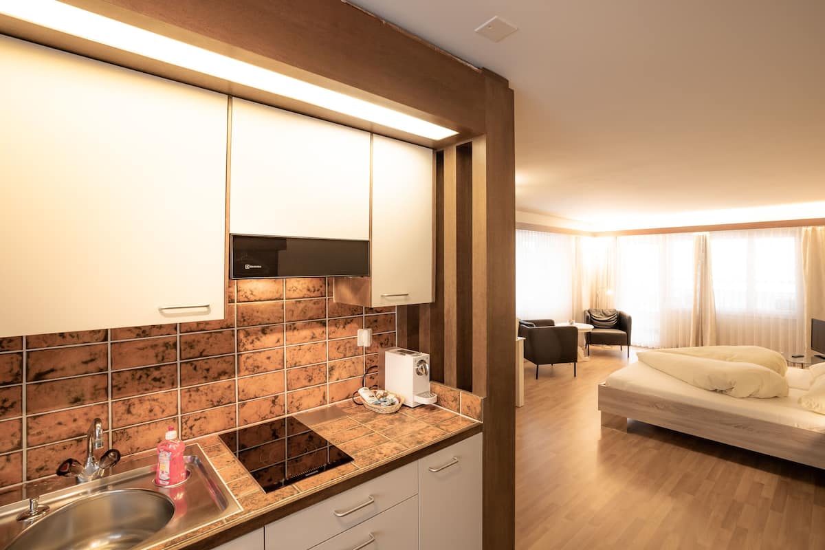 A modern hotel room with a kitchen area, a bed, a seating area, and a TV.
