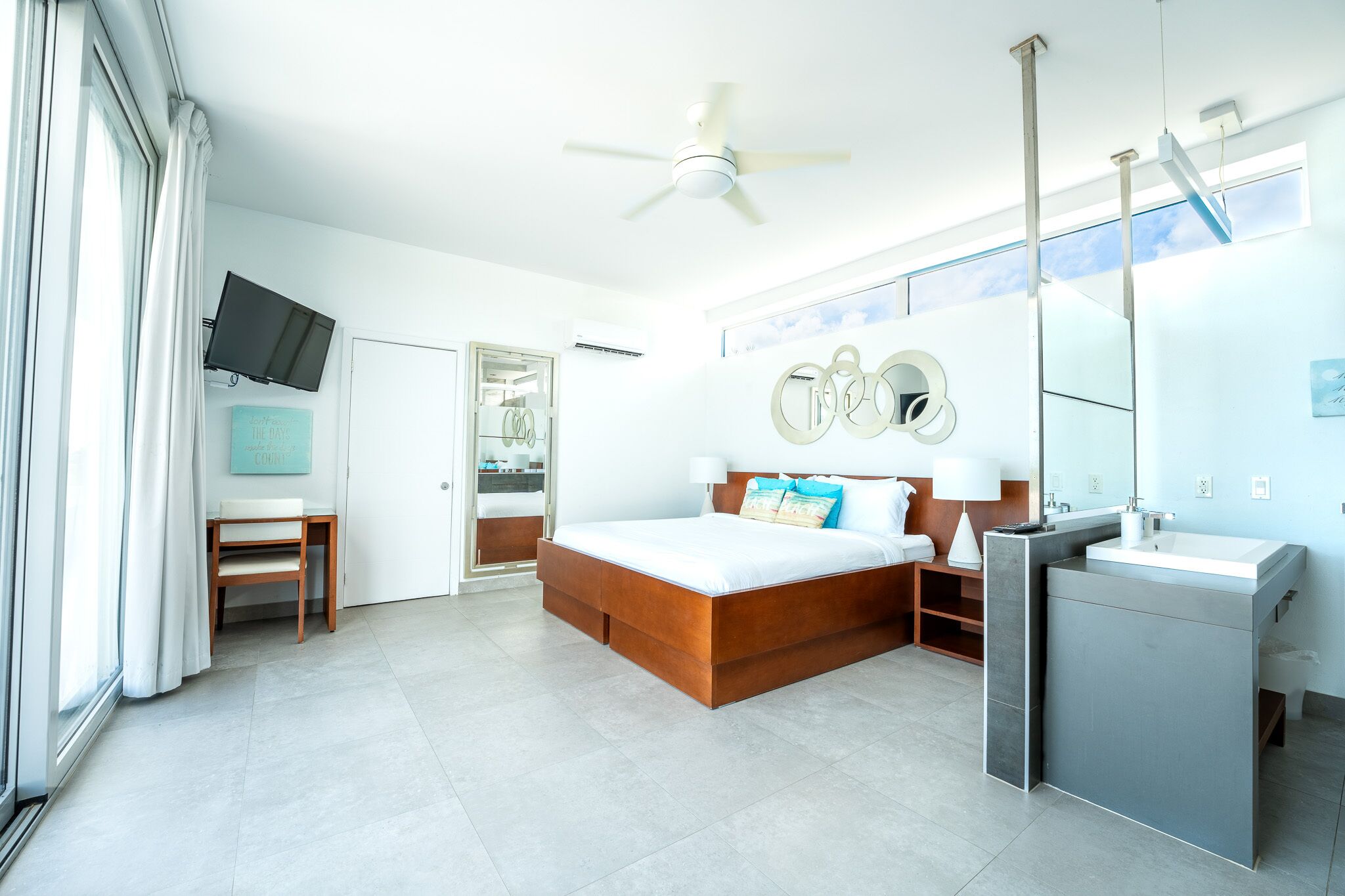A modern bedroom with a large bed, a built-in vanity, a ceiling fan, and a TV mounted on the wall.