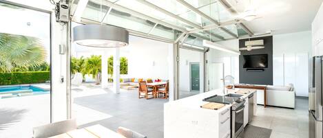 A modern kitchen with a central island, a dining area with a glass enclosure, and a pool visible through the glass doors.