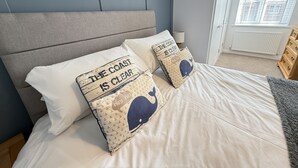 Quarto com cama, duas almofadas com motivos náuticos, cabeceira cinza, janela com persianas e radiador.