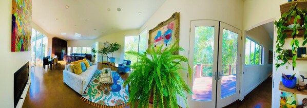 A spacious living area with a sofa, a coffee table, and a large fern plant. There is a painting on the wall and a door leading to an outdoor deck.