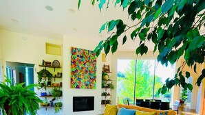 Een moderne woonkamer met een bank, salontafel en potplanten. Er is een open haard en een groot raam met uitzicht op groen.